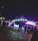 A large illuminated globe with the word 'Universal' is prominent at the center, surrounded by a misty, atmospheric effect. People are walking around the area, with some standing and looking at the globe. In the background, there are bright electronic billboards and palm trees, contributing to a lively and bustling environment.
