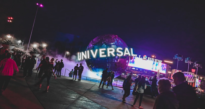 A large illuminated globe with the word 'Universal' is prominent at the center, surrounded by a misty, atmospheric effect. People are walking around the area, with some standing and looking at the globe. In the background, there are bright electronic billboards and palm trees, contributing to a lively and bustling environment.
