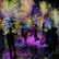 A vibrant and immersive digital art installation with bright, colorful light projections resembling flowers and abstract patterns covering the walls and floor. People are interacting with the environment, some sitting and others standing or walking around, creating an interactive and dynamic atmosphere.