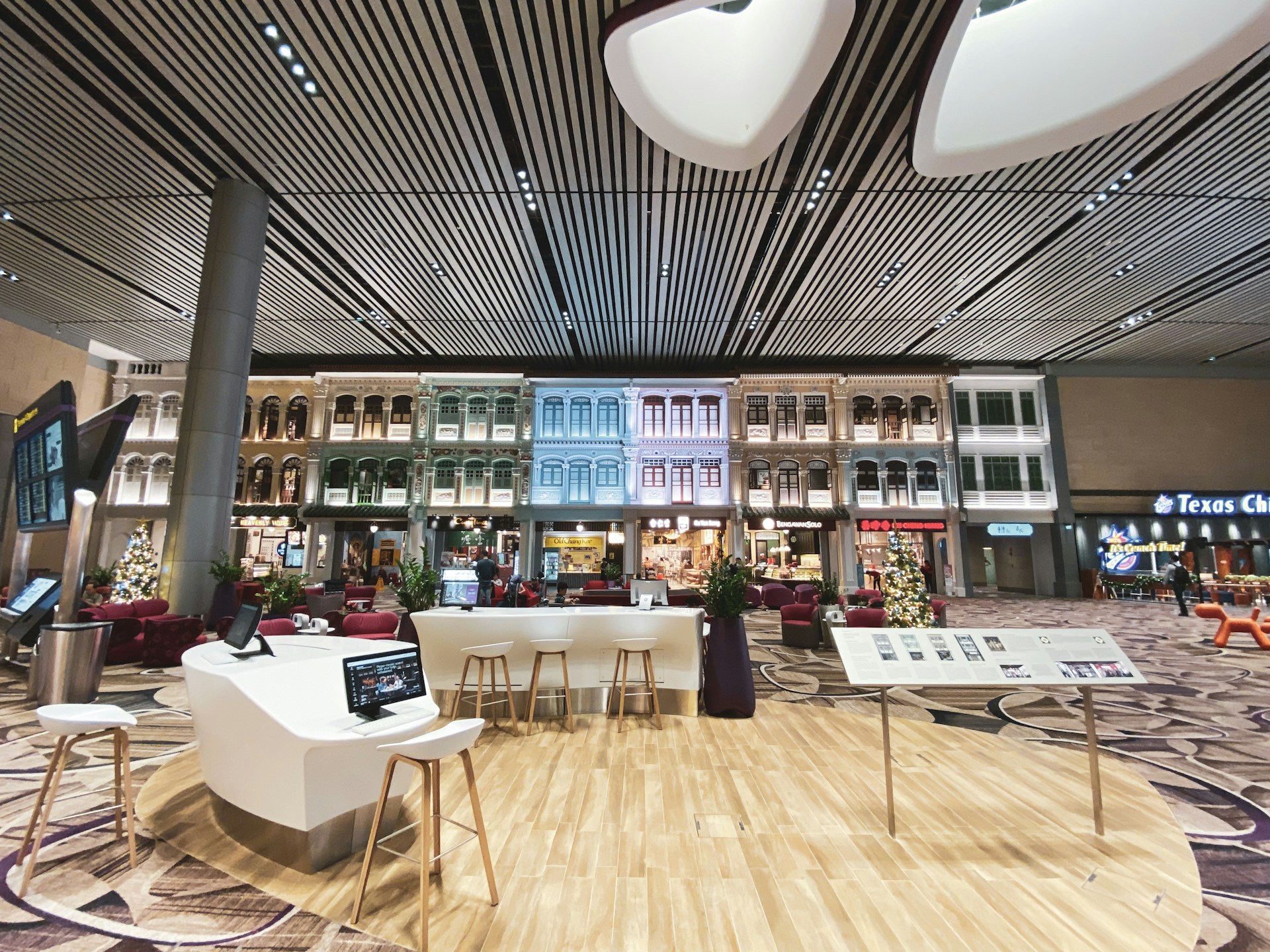 An indoor setting with a modern aesthetic, featuring a high, striped ceiling and walls that are designed to resemble traditional exterior building facades. The space includes contemporary seating arrangements, interactive screens, and digital kiosks. Christmas decorations, including a tree, are visible, suggesting a festive atmosphere.