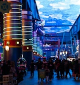 A vibrant indoor complex with illuminated columns and a large digital ceiling display showing a sky pattern. The area is bustling with people, possibly visiting an amusement park or an entertainment venue. There are colorful lights and signage directing to different attractions.