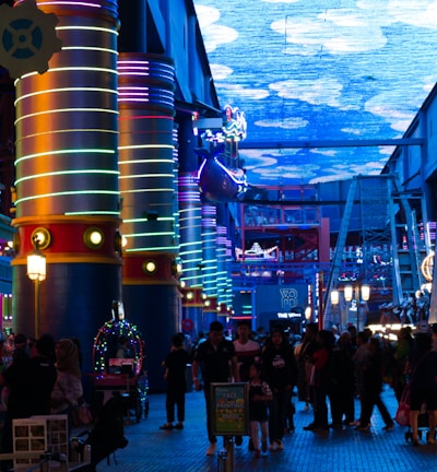 A vibrant indoor complex with illuminated columns and a large digital ceiling display showing a sky pattern. The area is bustling with people, possibly visiting an amusement park or an entertainment venue. There are colorful lights and signage directing to different attractions.