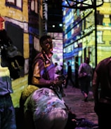 People are standing and walking in a dimly lit room with colorful digital projections on the walls, creating an immersive art experience. The projections depict a detailed building facade. The atmosphere appears lively with several individuals engaging with the artwork.