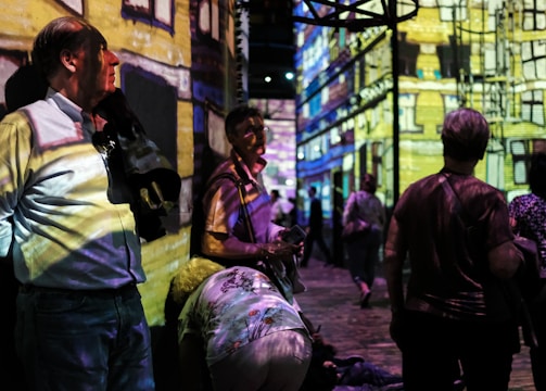 People are standing and walking in a dimly lit room with colorful digital projections on the walls, creating an immersive art experience. The projections depict a detailed building facade. The atmosphere appears lively with several individuals engaging with the artwork.