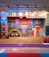A colorful and vibrant stage design featuring geometric patterns and abstract shapes. The central figure has a stylized face with a hat, surrounded by various symmetrical designs in bright colors. The stage is lit with spotlights and decorated with a banner that reads 'EMJOY Empower the Youth's Nextperience'.
