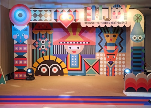 A colorful and vibrant stage design featuring geometric patterns and abstract shapes. The central figure has a stylized face with a hat, surrounded by various symmetrical designs in bright colors. The stage is lit with spotlights and decorated with a banner that reads 'EMJOY Empower the Youth's Nextperience'.