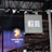 Hanging signage with bold, stylized lettering is displayed in a large indoor venue. Below, a digital display showcases a logo featuring a lion's head and text with vibrant blue and red lighting effects. The ceiling is lined with intricate lighting fixtures and metal trusses.