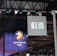 Hanging signage with bold, stylized lettering is displayed in a large indoor venue. Below, a digital display showcases a logo featuring a lion's head and text with vibrant blue and red lighting effects. The ceiling is lined with intricate lighting fixtures and metal trusses.