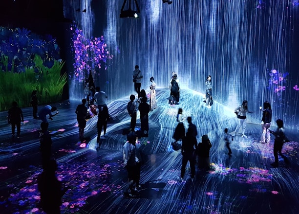 A group of people are standing and walking inside an immersive art installation with flowing digital streams and vibrant floral projections on the walls and floor. The environment appears interactive, with the participants seemingly engaged with the digital elements around them.
