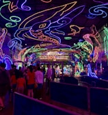 A vibrant and colorful space filled with neon light designs on the ceiling and walls, creating an immersive atmosphere. People are gathered in a queue, likely waiting to enter an attraction. The environment is reminiscent of an aquarium or theme park setting, with signs and decorations enhancing the lively mood.