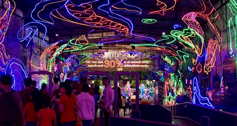 A vibrant and colorful space filled with neon light designs on the ceiling and walls, creating an immersive atmosphere. People are gathered in a queue, likely waiting to enter an attraction. The environment is reminiscent of an aquarium or theme park setting, with signs and decorations enhancing the lively mood.