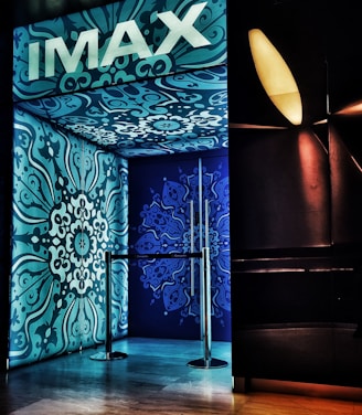 An entrance with ornate blue and teal designs covers the walls and ceiling, featuring intricate floral patterns. The brand 'IMAX' is prominently displayed at the top in bold white letters. A metallic barrier is present at the entryway, and ambient lighting creates a modern cinematic ambiance.