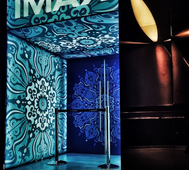 An entrance with ornate blue and teal designs covers the walls and ceiling, featuring intricate floral patterns. The brand 'IMAX' is prominently displayed at the top in bold white letters. A metallic barrier is present at the entryway, and ambient lighting creates a modern cinematic ambiance.