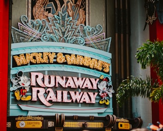 A colorful sign featuring playful typography and animated figures associated with a popular entertainment franchise. The sign is adorned with neon lights and decorative elements, set against a backdrop of bright red pillars and greenery.