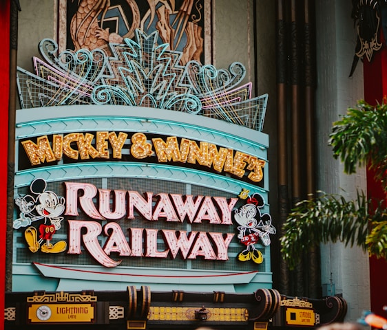 A colorful sign featuring playful typography and animated figures associated with a popular entertainment franchise. The sign is adorned with neon lights and decorative elements, set against a backdrop of bright red pillars and greenery.