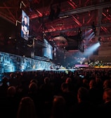 A large indoor event with a stage featuring bright screens and spotlights, attended by a substantial crowd. The venue has a high ceiling with industrial beams and red lighting accents. The atmosphere resembles a concert or a conference with a focus on visual effects.