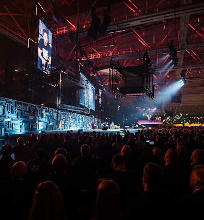 A large indoor event with a stage featuring bright screens and spotlights, attended by a substantial crowd. The venue has a high ceiling with industrial beams and red lighting accents. The atmosphere resembles a concert or a conference with a focus on visual effects.