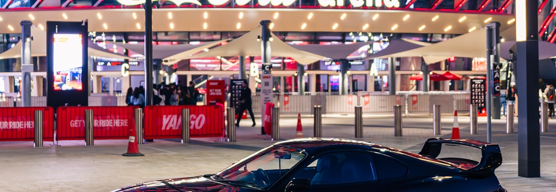 A sleek black sports car is parked in front of an illuminated arena entrance, with a large sign bearing the name of a famous beverage brand. The building features a modern, geometric facade with bright red and blue lights, creating a vibrant and lively atmosphere.